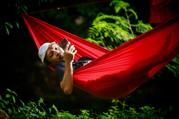 asian old man relaxing in hammock and looking to his mobile phone in the morning