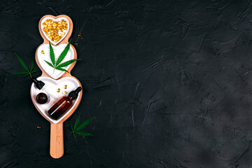 Heart dish with glass bottles with cbd oil, capsules and hemp leaves on black background. Top view, flat lay, copy space.