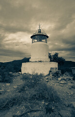 Fototapeta na wymiar View on the old white lighthouse of Monte Poro under dramatic sky, Elba island, Italy. Old abandoned lighthouse in the middle of the bushes. Vertical image, split tones.