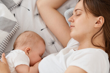 Obraz na płótnie Canvas Portrait of sleepy woman feeding breast her baby in the bed sleeping together, young adult female mommy wearing white casual t shirt, resting while her toddler kid sleeps.