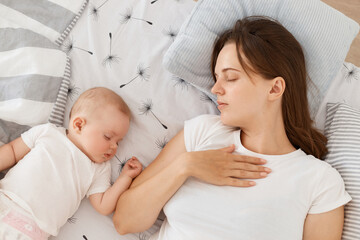 Young mother and her baby sleeping together in bed, keeps eyes closed, wearing white casual style t shirt, lying on pillow, tired mother resting while her cute toddler daughter sleeps.