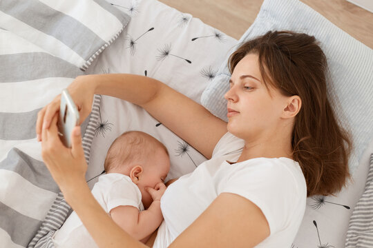 Good looking tired sleepy woman wearing white casual style t shirt using cell phone for checking networks or typing messages while breast feeding her infant baby, lying bed.