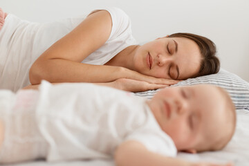 Fototapeta na wymiar Side view portrait of sleeping mother and toddler daughter lying on bed, family wearing white clothing, resting, early morning, motherhood and childhood.