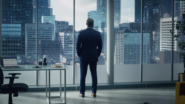 Thoughtful Black Businessman in a Tailored Suit Standing in Office Looking out of the Window on Big City. Successful Corporate Executive Thinking Investment Strategy for e-Commerce Startup. Back View