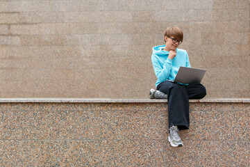 Young hipster woman with short hair sits with a laptop in an urban environment. She works hard, sitting on the parapet against the granite wall.