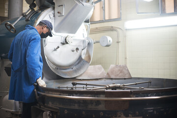 Worker in uniform controlling coffee roasting process. Coffee roaster working on roasting equipment. Man in mask and uniform Working with machinery appliance. Production, food concept