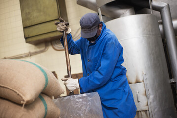 Man in uniform getting coffee ready for roasting. Coffee roaster working on roasting equipment. Man holding shovel. Man in mask and uniform Working with machinery appliance. Production, food concept