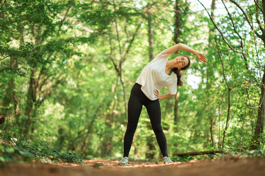 A young woman in sports clothes does a stretching exercise. Summer forest in the background. Low angle. The concept of training in the park