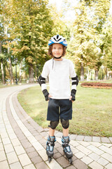 Vertical full length shot of a charming boy wearing rollerskates, protective skating pads and helmet at the park