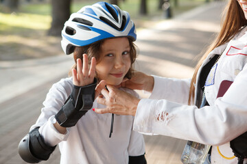 Cute boy waving to the camera, while his mother putting on skating helmet on him at the park © Ihor