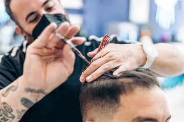 Barber with mask cutting the hair of a client using scissors in a barber's shop