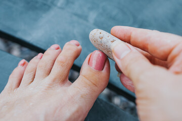 The girl on the beach smooths and polishes her toenail with an abrasive stone of pebbles - independent pedicure at the sea