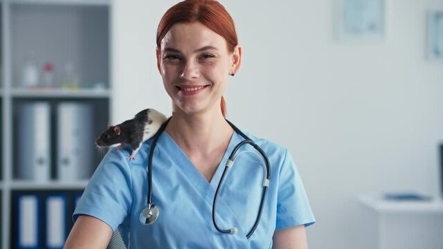 animal treatment, portrait of young charming female veterinary doctor with domestic rat on her shoulder smiling and looking at camera in medical office