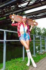 Caucasian Hipster Skateboarder Girl Posing With Longboard Under Rusty Metal Construction Outdoor While Holding  Skateboard Behind