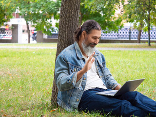 Middle aged man during online video call - he is sitting on the grass in city park with laptop. He waving hand - hello.