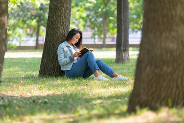 Asian woman reading a book in the park. She sitting on the grass under the tree, reads a book and...