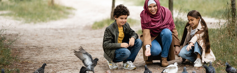 Muslim mother and kids feeding birds in park, banner