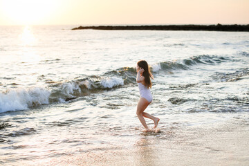 Fototapeta na wymiar Cute caucasian girl in a striped swimsuit with long hair runs seashore on a deserted beach at a resort, evening and sunset