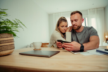 Caucasian couple sitting at kitchen table making online payment holding cellular device