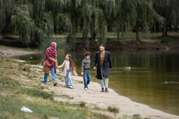 Fototapeta premium Positive arabian family holding hands while walking near lake