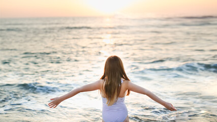 Cute caucasian child girl in a striped swimsuit with long hair with outstretched arms hugs the sea on an empty beach at a resort, evening and sunset