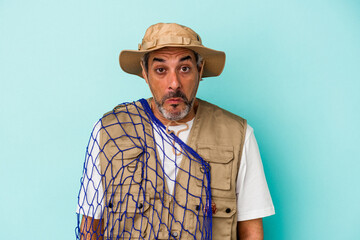 Middle age caucasian fisherman holding net isolated on blue background  shrugs shoulders and open...