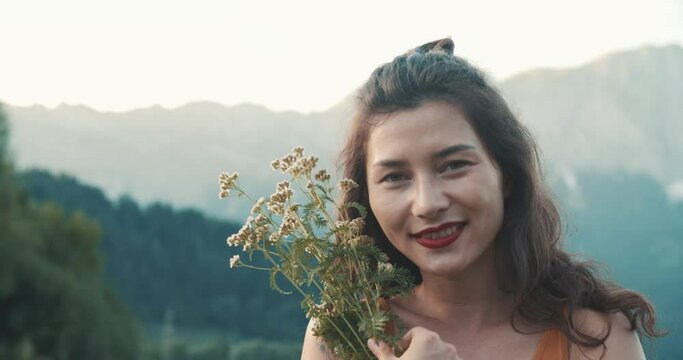 Cute Asian young woman with field flowers on mountain top above sea. Summer nature landscape. Girl tourist standing on cliff at seaside. The mood of cinematography, gentle tones, alpine meadows and