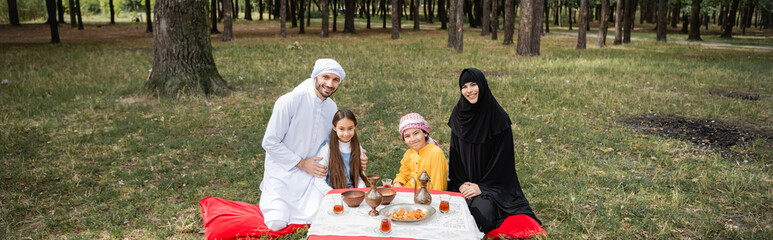 Smiling muslim family looking at camera near food in park, banner