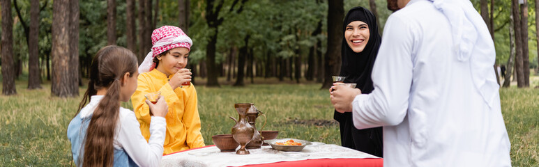 Smiling arabian woman sitting near husband, kids and tea in park, banner