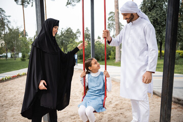 Smiling arabian parents looking at daughter on swing in park