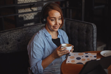 a middle-aged woman in a blue shirt undergoes online training on a macbook, sitting in headphones in a cafe over a mug of coffee and makes notes in a notebook