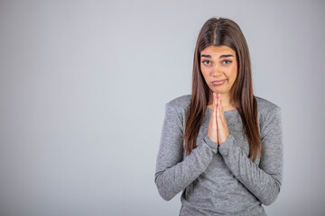 Young  woman pleading and asking for something with clasped hands, positive emotional lady looking at camera with please expression, isolated over gray background