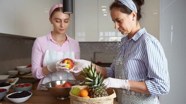 Attractive caucasian women in aprons and gloves sorting fruits from basket while standing on modern kitchen. Two females choosing only fresh and organic ingredients for healthy eating.