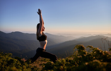 Side view of sporty young woman doing yoga exercise on grassy hill with blue morning sky on background. Fit woman in sportswear working out outdoors in mountains.