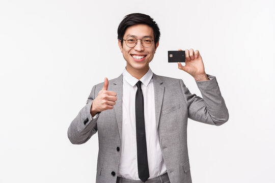 Waist-up Portrait Of Satisfied Good-looking Asian Businessman Using Banking Service, Keep His Money Safe On Bank Account, Show Credit Card, Smiling And Showing Thumbs-up, White Background