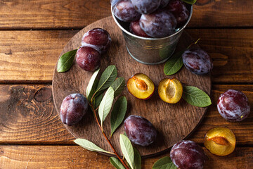Ripe plums on a wooden cutting board, among green leaves and twigs. One plum is cut in half and the pulp and bone are visible.