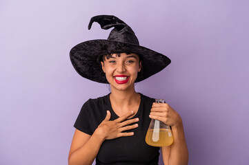 Young mixed race woman disguised as a witch holding potion isolated on purple background  laughs out loudly keeping hand on chest.