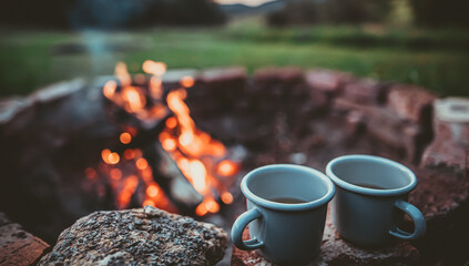 Campsite With Fire Pit and Two Tin Cups with hot tea. Burning Campfire with mountain landscape with...