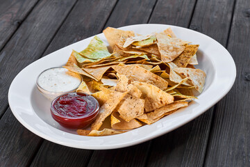 Triangular nachos chips with tomato ketchup and cheese sauce on a white plate and wooden background