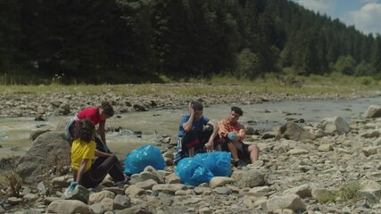 Group of tired positive diverse multiracial eco activists catching breath, talking and resting at mountain riverbank on hot summer day after successful environmental cleanup in mountains.