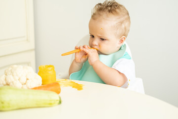 funny baby boy in bib eating vegetable puree with spoon sitting in child chair. Child learn to eat by himself