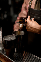 hand of male bartender masterfully pours drink from jigger into steel shaker glass