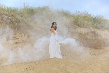 Fototapeta na wymiar Junge schöne Frau im Hochzeitskleid am Strand mit einer Rauchfackel und weißen Rauch