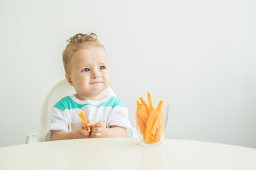 Baby boy sitting in a Childs chair eating carrot slices on white background.