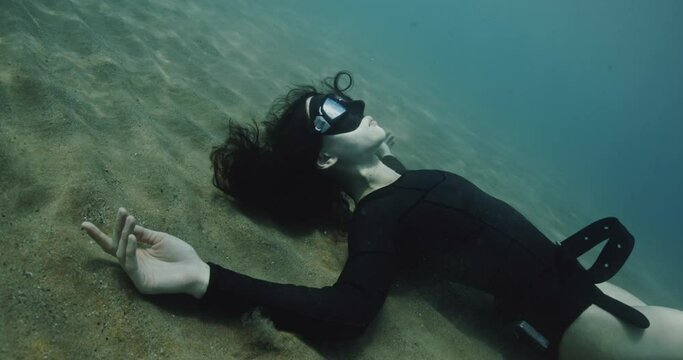 Diver woman lying underwater bottom, filmed on cinema camera. Brutal girl in black suits against background of mountains and ocean. Beautiful brunette touching her shoulder. Concept of arthouse and