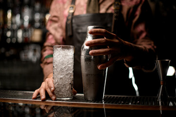 Close-up of transparent glass with crushed ice and a steel shaker standing on the bar