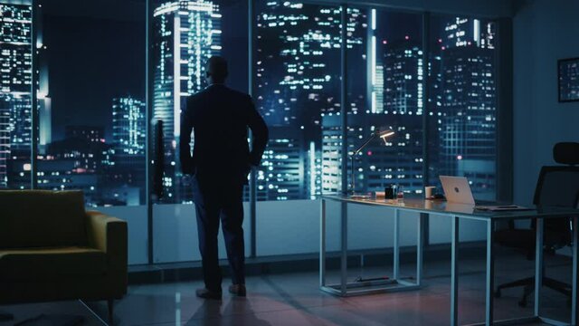 Night Office: Successful Black Businessman Wearing Suit Standing, Looking out of the Window on a Big City. African-American CEO Thinking of Investment Strategy in e-Commerce Startup