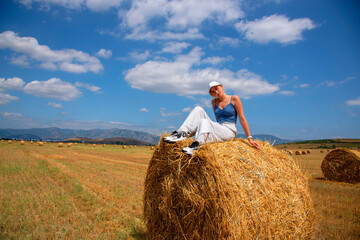 Naklejka na ściany i meble A girl is sitting on a haystack in a huge field against the background of mountains on a sunny August day.
