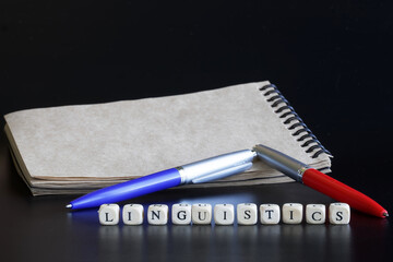 Notepad, two pens and the word linguistics, made up of wooden cubes on a dark background. A concept...