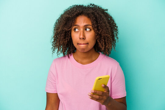 Young African American Woman Holding A Mobile Phone Isolated On Blue Background Confused, Feels Doubtful And Unsure.